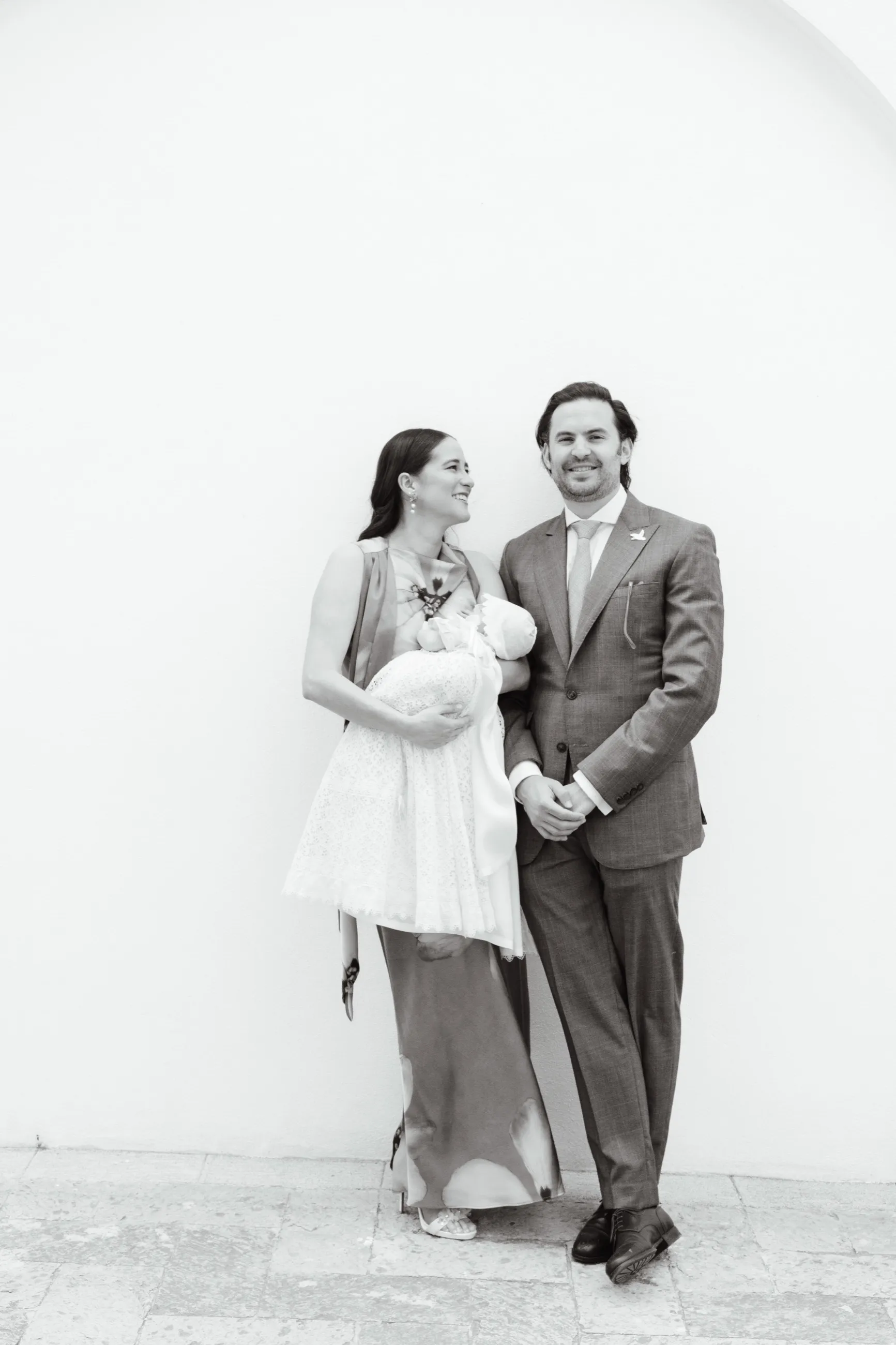 Pareja con un bebé en brazos vestido con ropón de bautizo, ante una pared blanca, en blanco y negro
