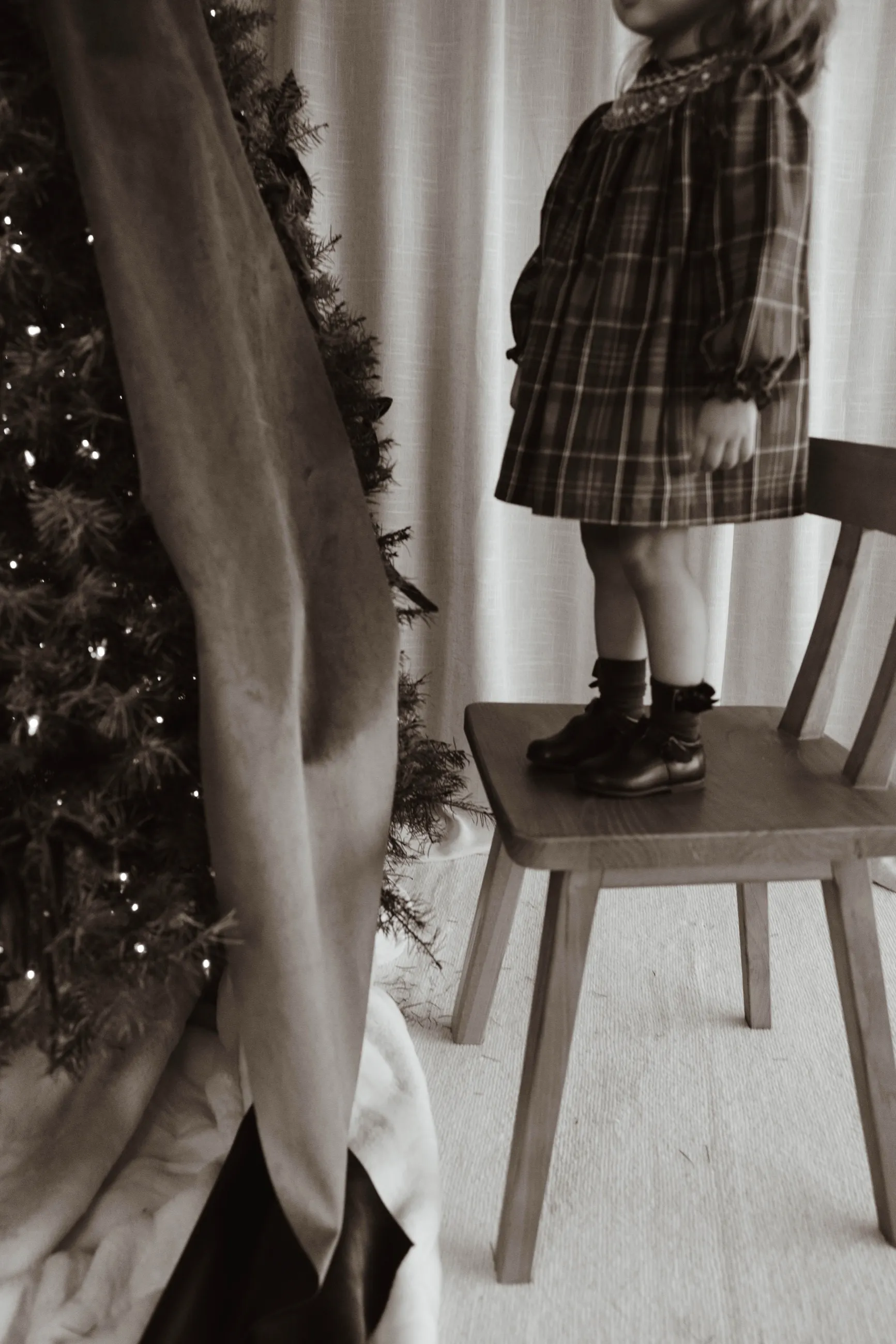 Niña con vestido de cuadros de pie sobre una silla junto a un árbol de Navidad iluminado, en blanco y negro