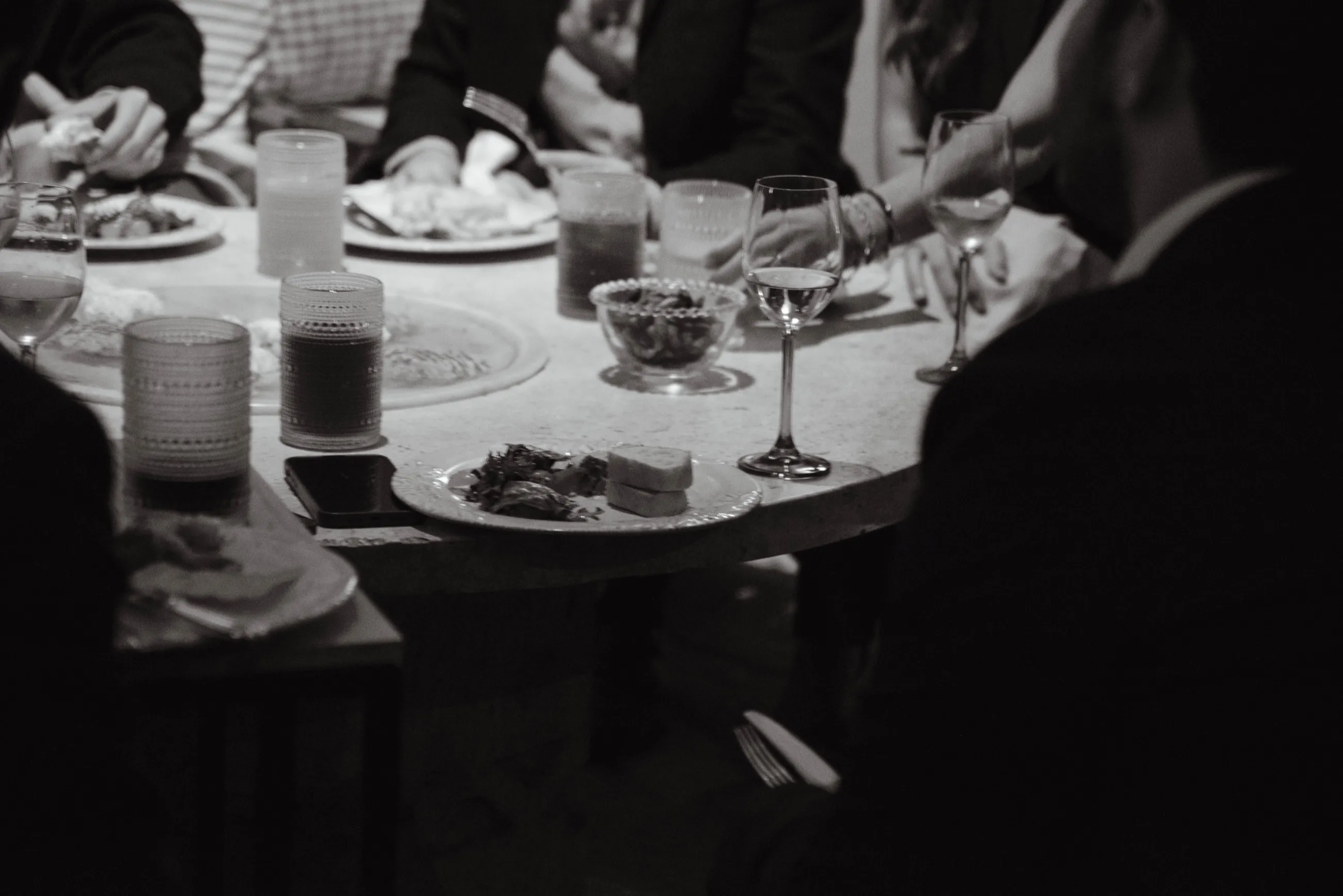 Mesa de cena durante un evento, con copas e invitados alrededor, en blanco y negro