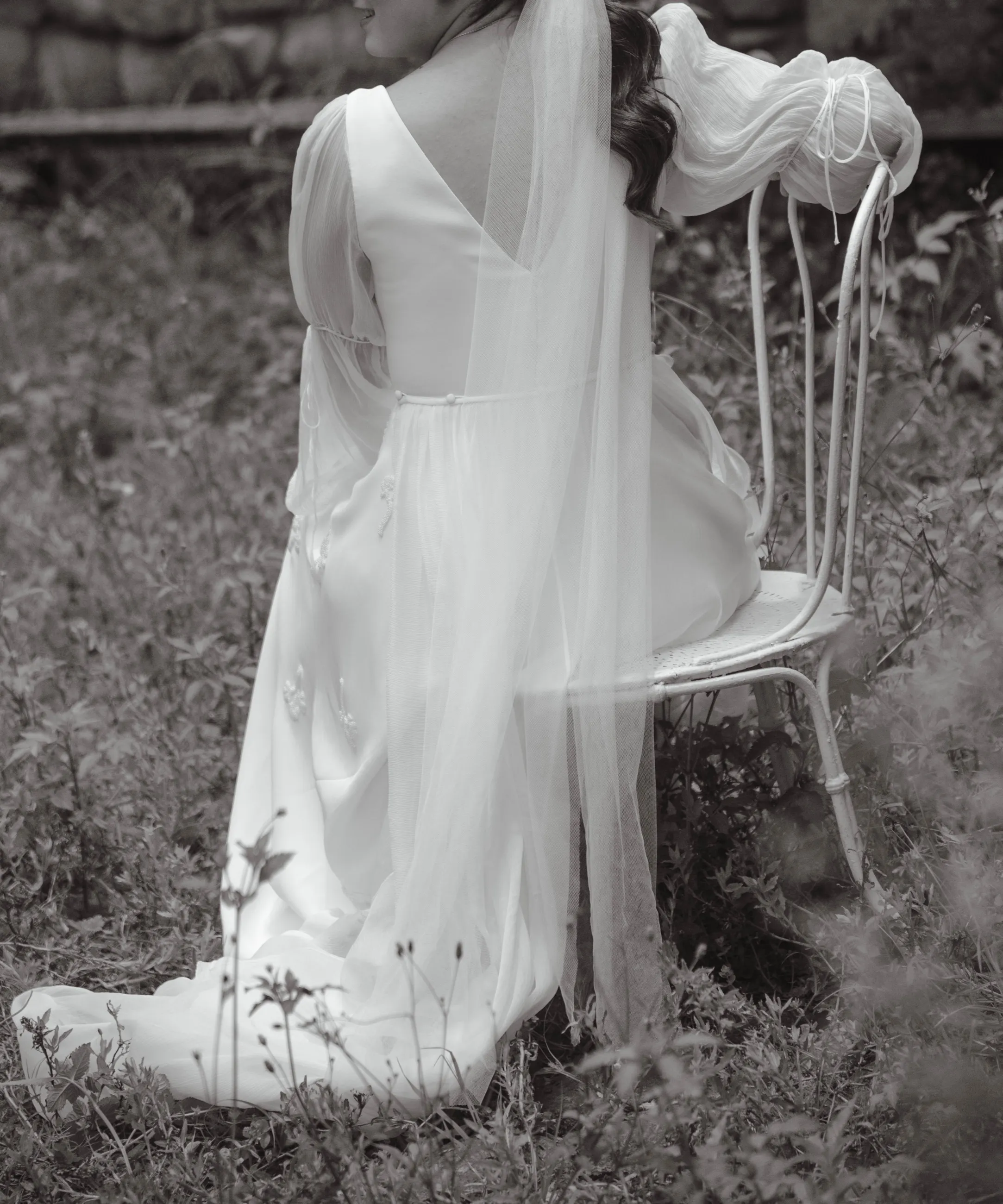 Novia de espaldas con vestido de tul y cola larga apoyada en una silla en un jardín, en blanco y negro