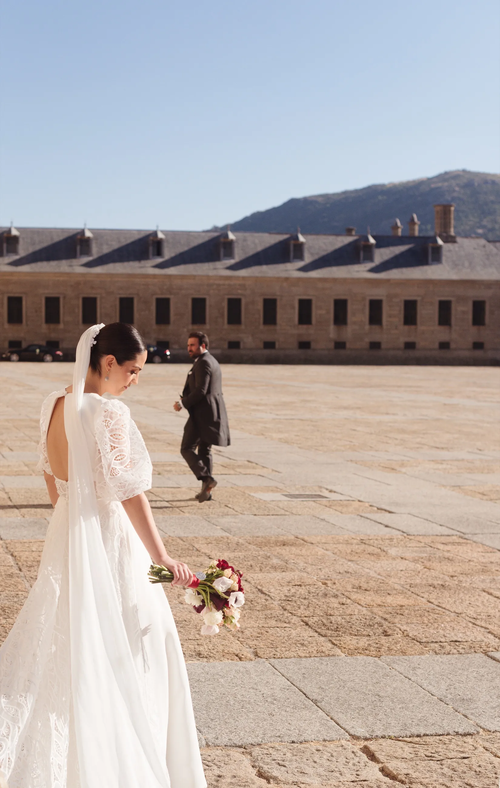 Novia con velo y ramo en primer plano y novio caminando al fondo por una explanada empedrada frente a un edificio monumental de piedra